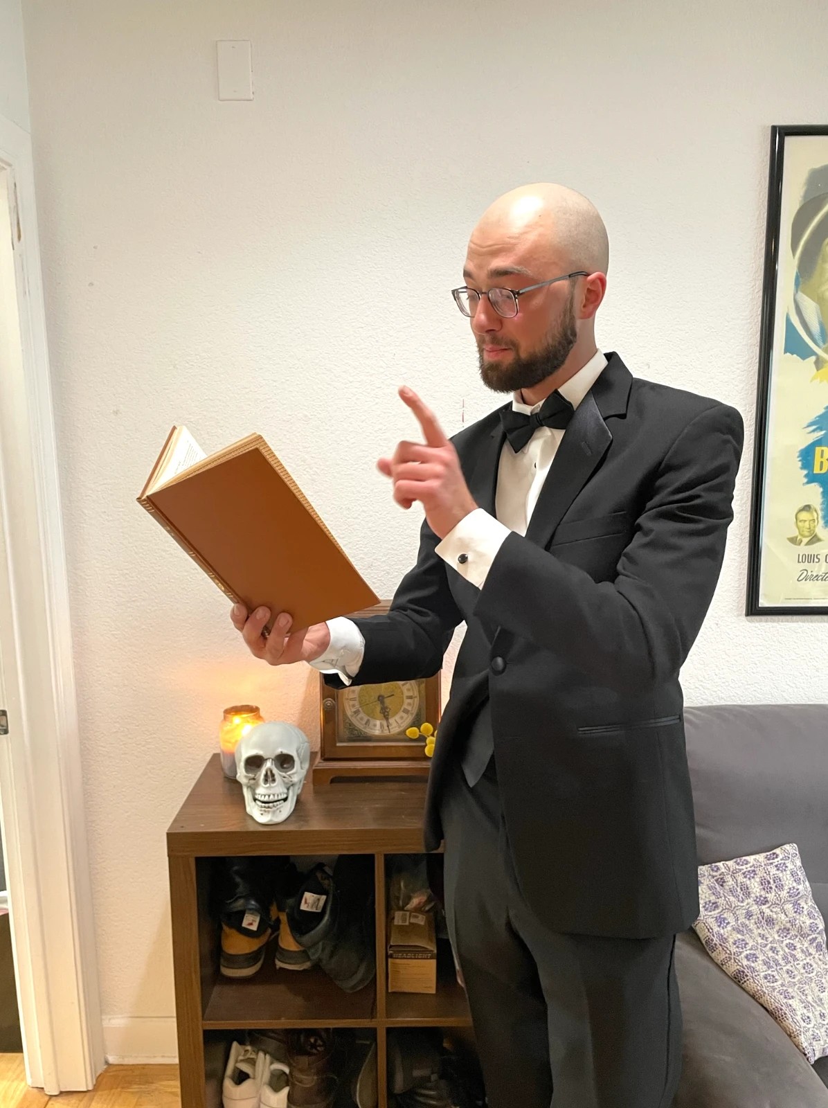 Carter in a tuxedo reading a book and pointing, beside a skull and an antique clock